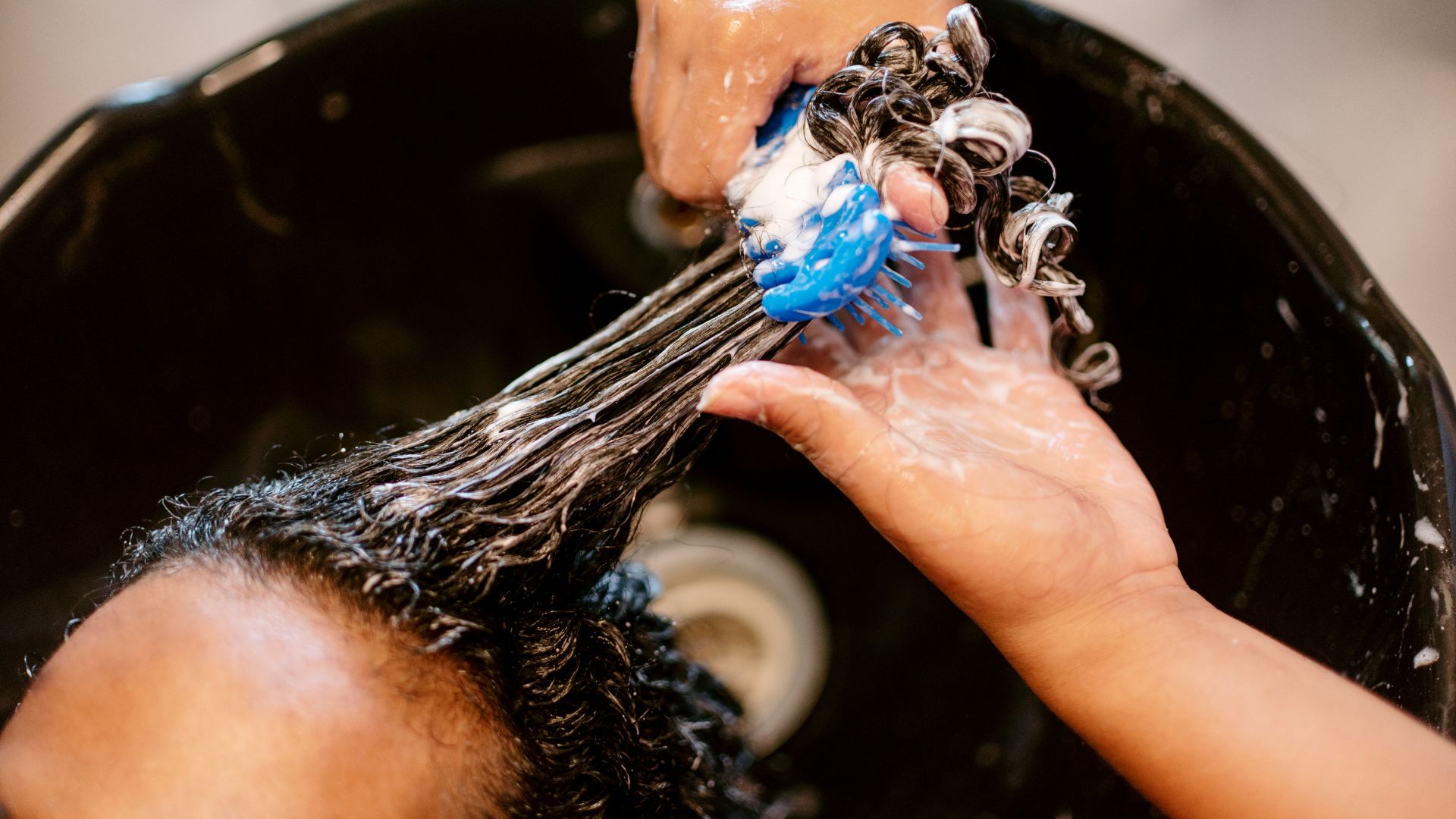 Hands washing curly hair with blue brush in a black sink